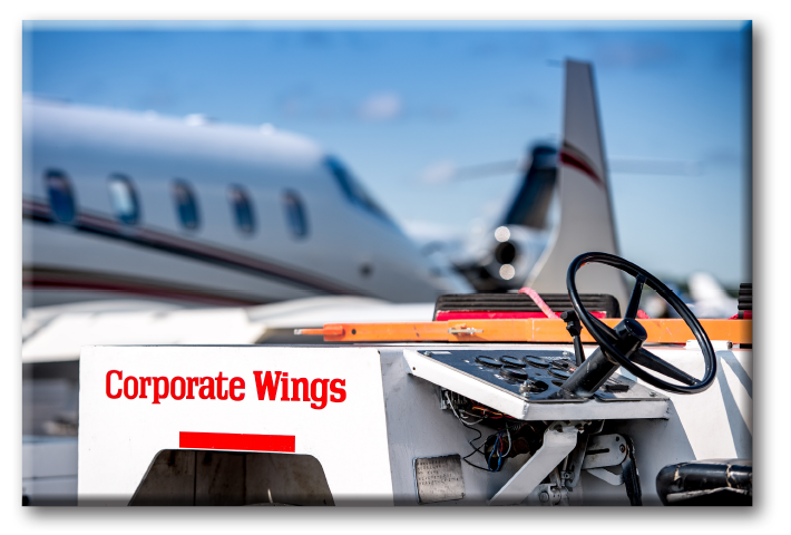 Outdoor image of a private airport tarmac, a private jet and an aircraft line-service tug with the name Corporate Wings on the side.